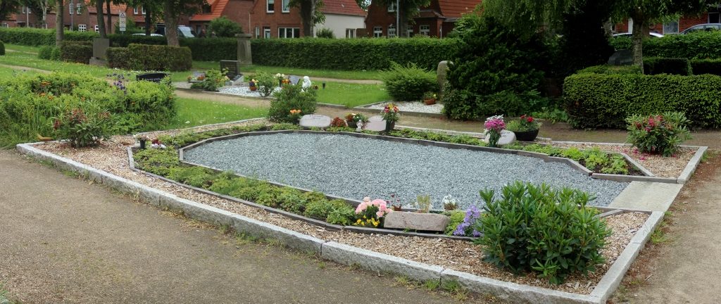 Ein Oval aus Kieselsteinen, rund herum bepflanzt und liegende Grabsteine.