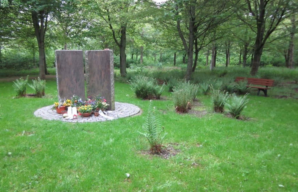 Zwei hohe Steine in einem Wald, darum eine runde Steinablagefläche mit Blumen.