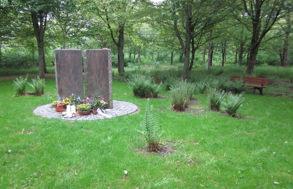 Zwei hohe Steine in einem Wald, darum eine runde Steinablagefläche mit Blumen.