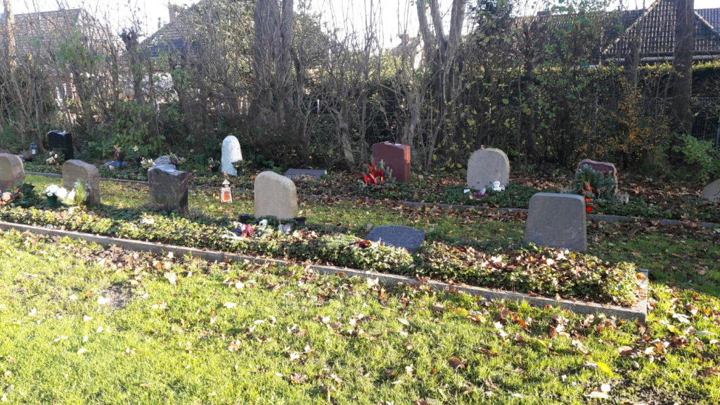Viele stehende Grabmale in zwei Reihen mit etwas Bepflanzung.